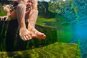 Person’s feet dangling over moss-covered stone steps under clear water in a natural swimming pond